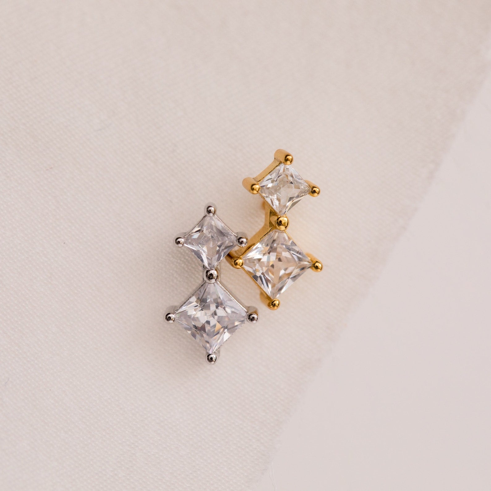 Aria Diamond Drop Studs featuring gold and silver crystal with three square gemstones, displayed on a light fabric background.