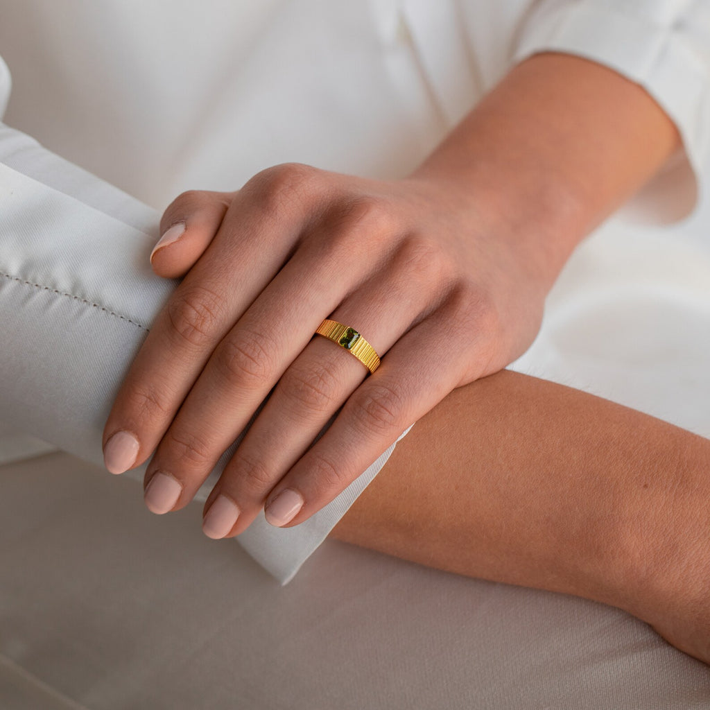 A hand with nude nails wearing the Olive Peridot Signet Ring rests gently on another person's arm in soft lighting.