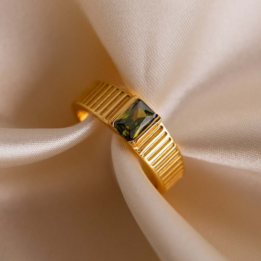 The Olive Peridot Signet Ring features a gold chunky, textured band with an olive peridot green gemstone, displayed on soft cream-colored fabric.