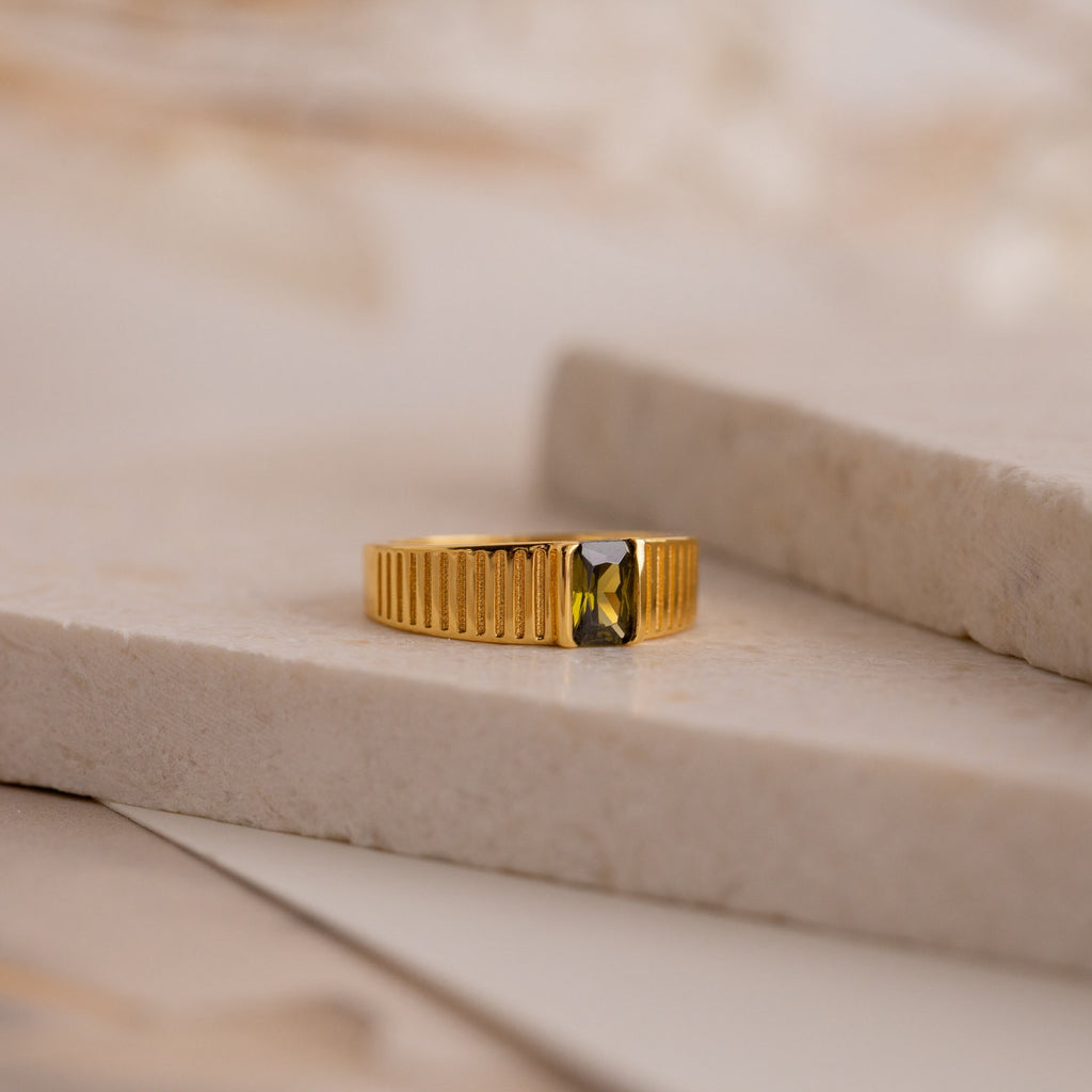 The Olive Peridot Signet Ring, featuring a bold rectangular green gemstone, rests elegantly on a marble surface.