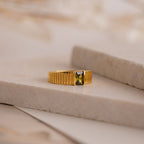 The Olive Peridot Signet Ring, featuring a bold rectangular green gemstone, rests elegantly on a marble surface.
