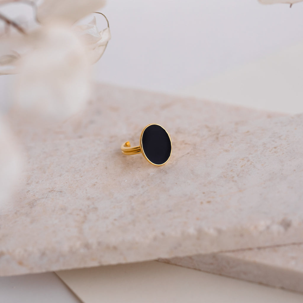 The Midnight Hour Onyx Ring features an oval black onyx stone set in gold, shown on a marble surface with softly blurred flowers in front. This vintage-glam ring offers an adjustable band for a comfortable fit.