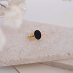 The Midnight Hour Onyx Ring features an oval black onyx stone set in gold, shown on a marble surface with softly blurred flowers in front. This vintage-glam ring offers an adjustable band for a comfortable fit.