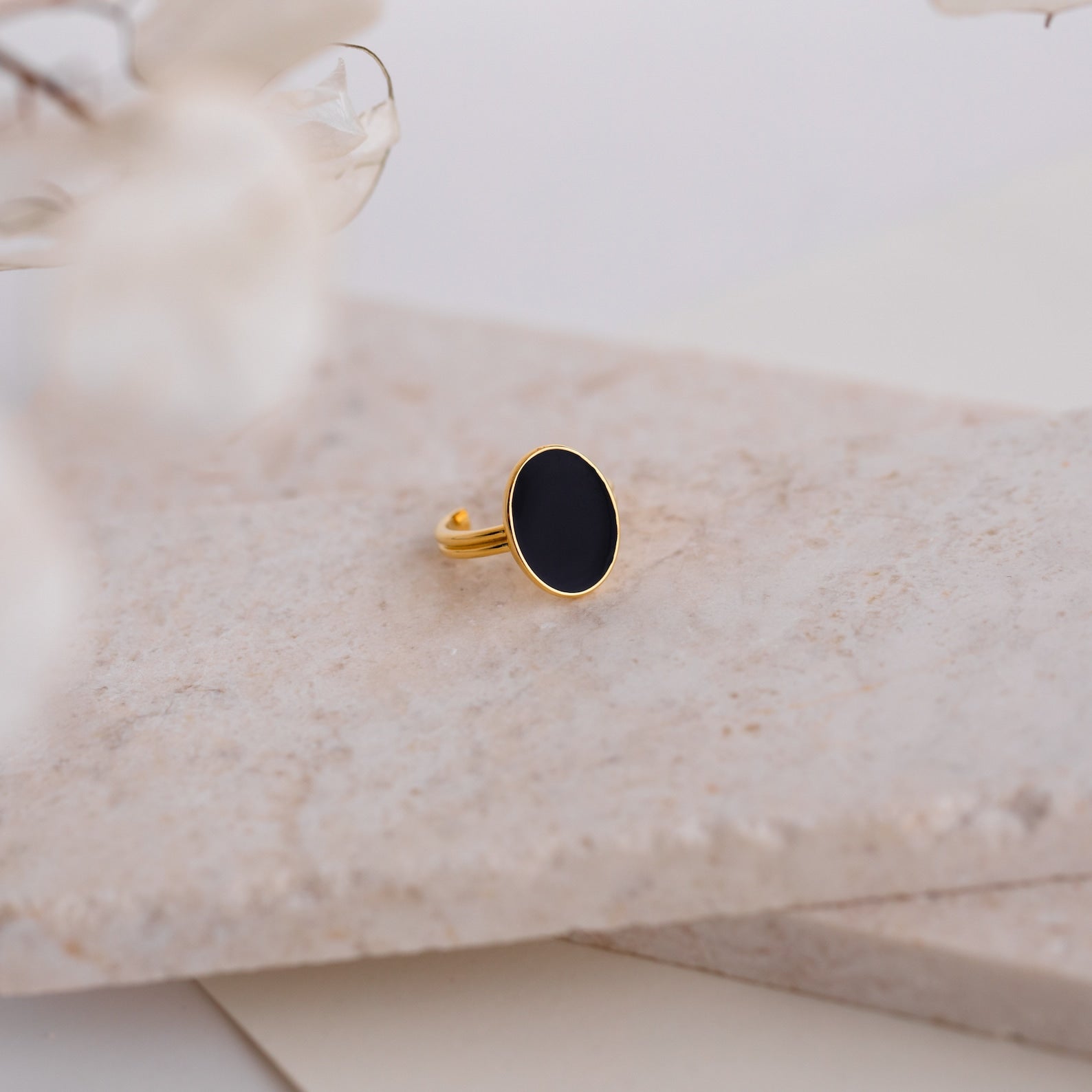 The Midnight Hour Onyx Ring features an oval black onyx stone set in gold, shown on a marble surface with softly blurred flowers in front. This vintage-glam ring offers an adjustable band for a comfortable fit.