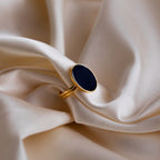 The Midnight Hour Onyx Ring, a gold signet style with a black oval stone, rests on smooth cream satin—a vintage-glam piece designed with an adjustable band for the perfect fit.