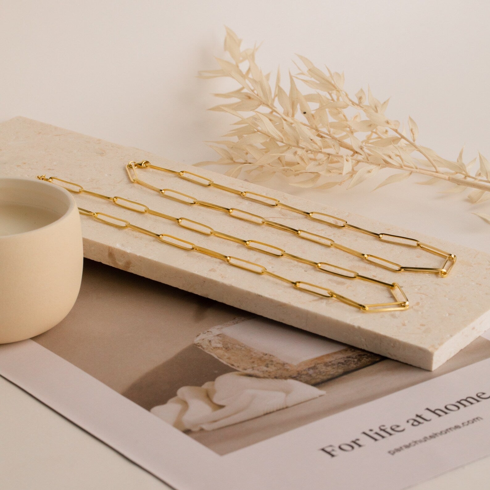 The Custom Link Chain Necklace sits on a marble slab beside a beige cup, dried leaves, and a magazine—an ideal bold accessory for making statement necklaces stand out.
