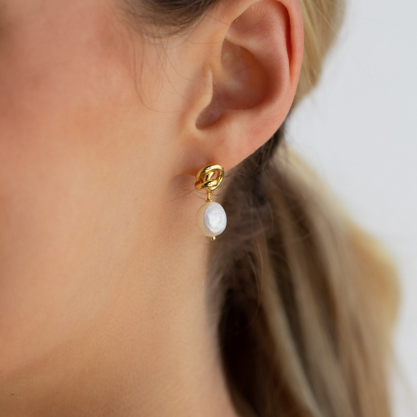 Close-up of Knot Pearl Drop Earrings, featuring gold and pearl accents, shown on someone with light brown hair.