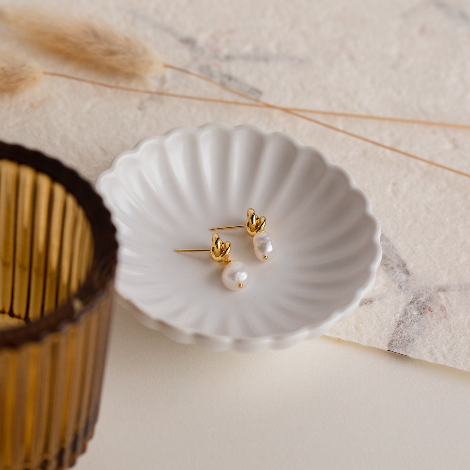Knot Pearl Drop Earrings in 18K Gold, featuring a gold Celtic knot and pearls, are displayed on a white scalloped dish with a brown glass and dried grass nearby.