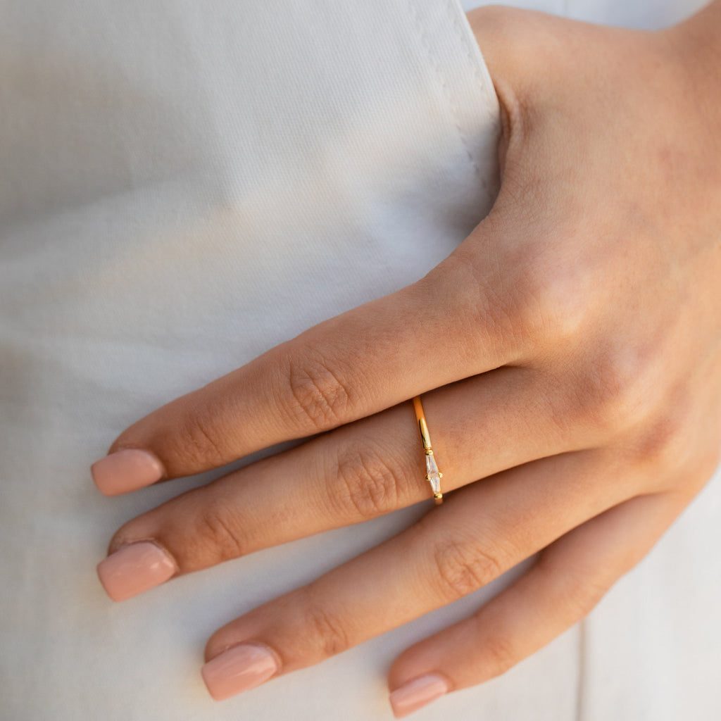 A hand with nude-polished nails wears the Eleanor Baguette Diamond Ring, which has a thin gold band set with a small rectangular diamond crystal baguette.