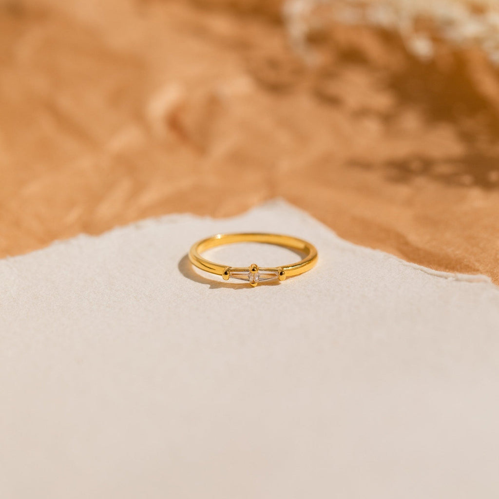 The Eleanor Baguette Diamond Ring features a delicate gold band with a small diamond baguette, displayed on a beige and tan textured background.