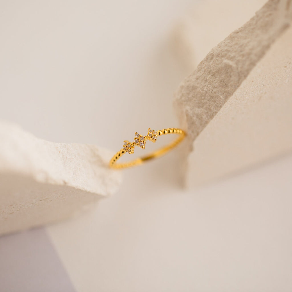 The Jasmine Beaded Diamond Ring features two small star-shaped charms in gold, ideal for stacking, and is showcased on a neutral, textured background.