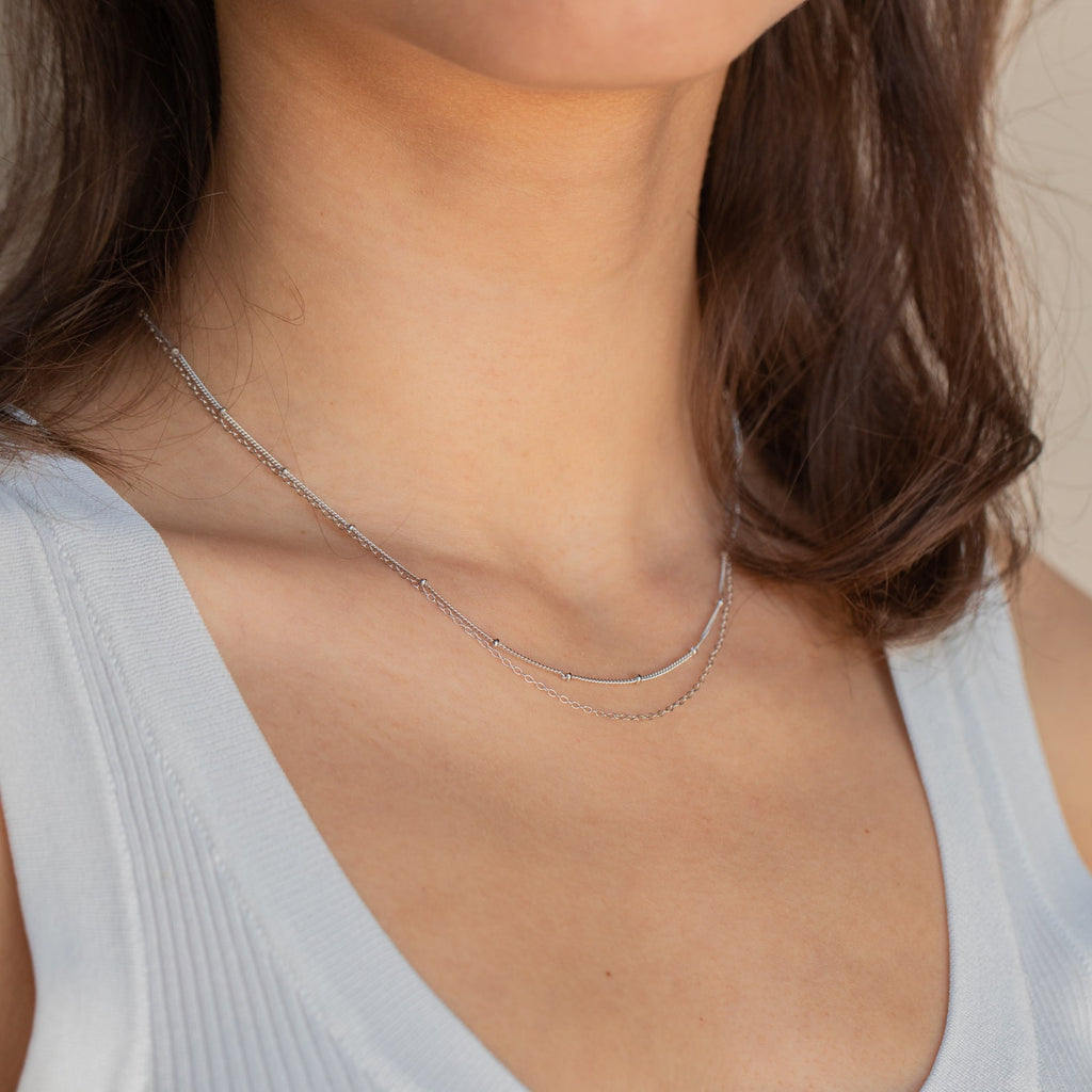 A woman elegantly pairs a light blue top with the Beaded Duo Chain Necklace, a minimalist double-layered silver chain.