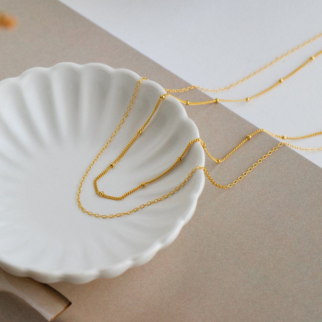 Two delicate gold necklaces, one with a simple cable chain and the other featuring small evenly spaced beads, draped across a white scallop-edged dish.