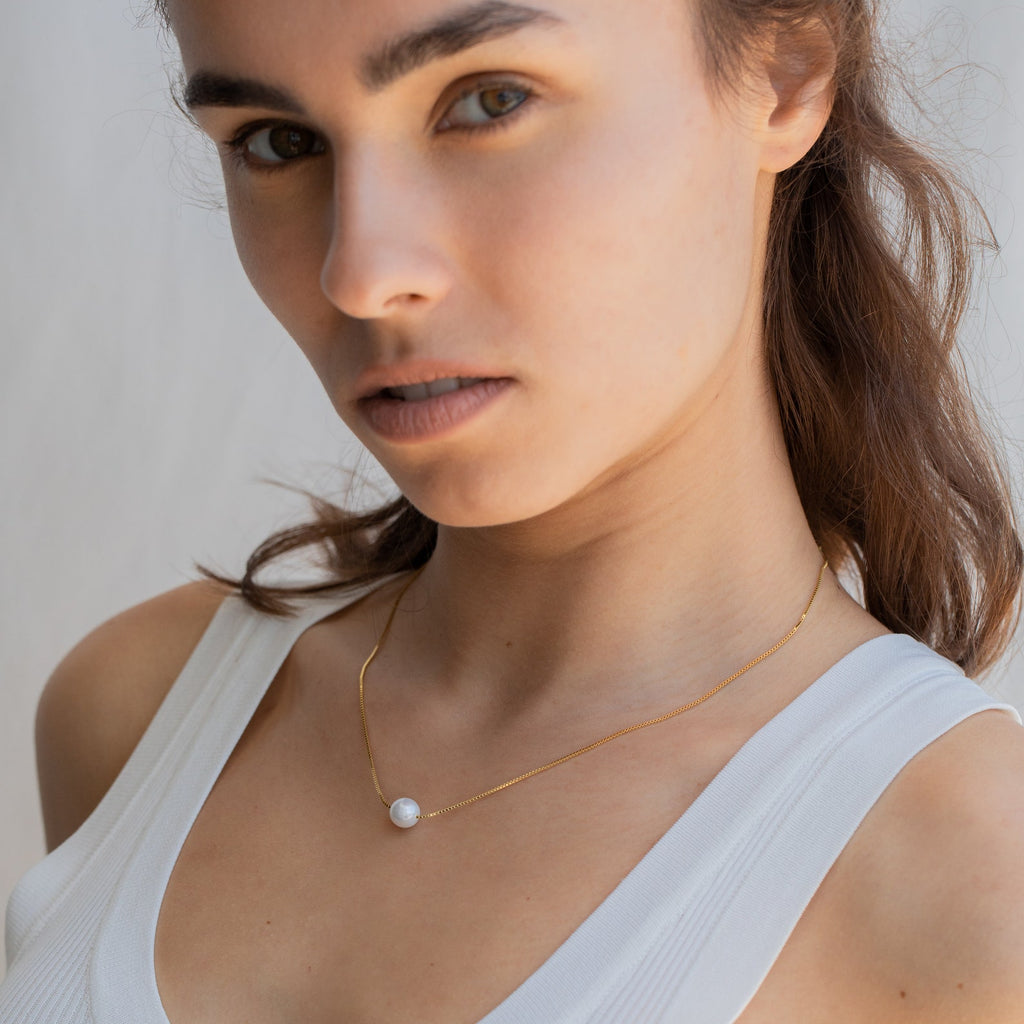 Woman in a white tank top wears the Kennedy Pearl Necklace, a gold pendant that adds minimalist elegance and sophistication to her look as she gazes at the camera.