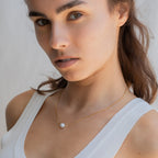 Woman in a white tank top wears the Kennedy Pearl Necklace, a gold pendant that adds minimalist elegance and sophistication to her look as she gazes at the camera.