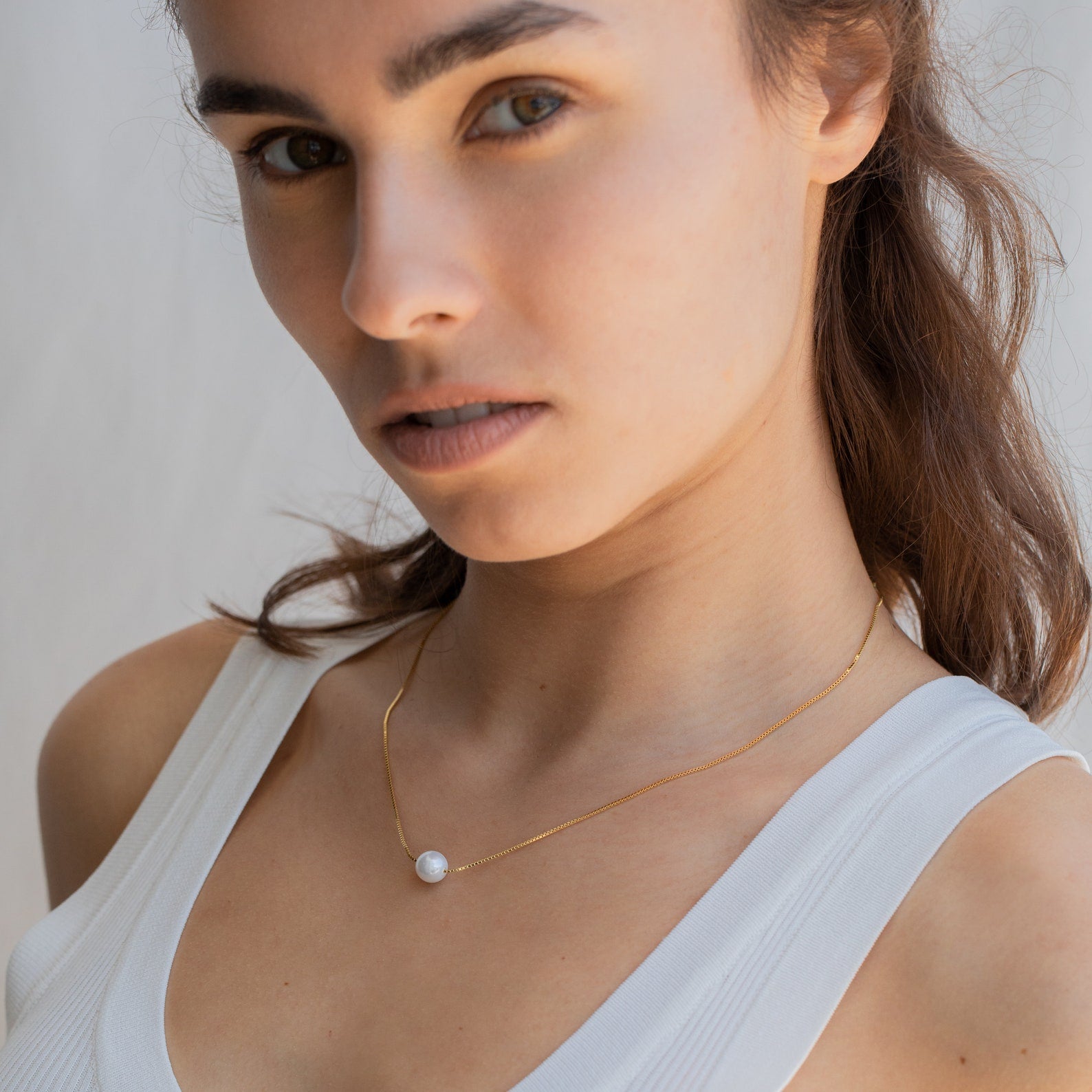 Woman in a white tank top wears the Kennedy Pearl Necklace, a gold pendant that adds minimalist elegance and sophistication to her look as she gazes at the camera.