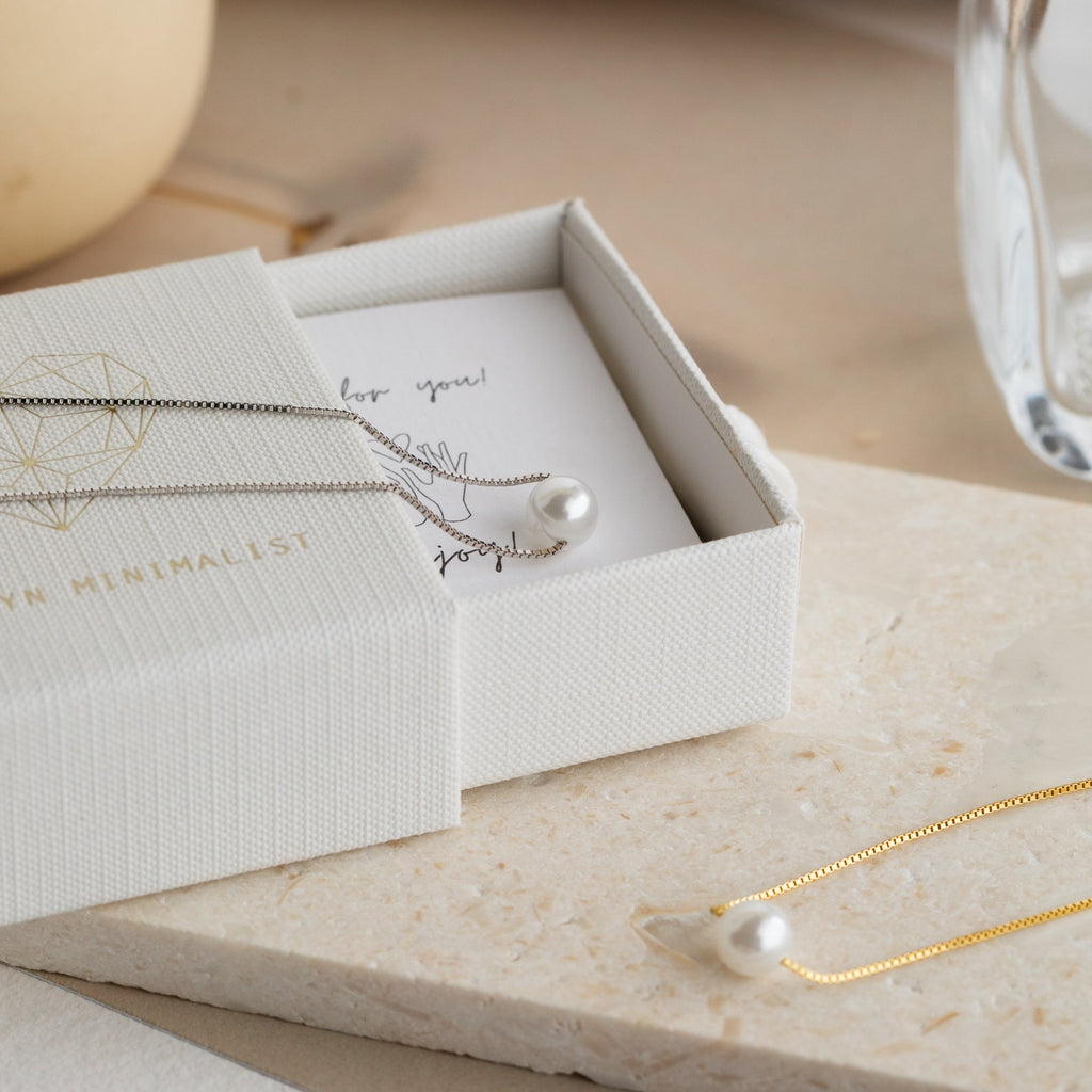 The Kennedy Pearl Necklace, a minimalist silver and gold pendant, is perfect for weddings or everyday wear and is elegantly displayed on a cream box and stone surface.