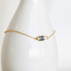 The Eloise Gemstone Bracelet features a delicate chain with a blue rectangular gemstone and two small white stones, highlighting dainty jewelry style, shown on a white vase.