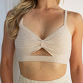 Model wearing a thin gold body chain with a Y shaped design, draping from the neck through the center of the torso and looping around the waist, paired with a cream ribbed bralette and high waisted bottoms.