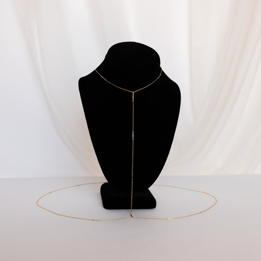 The Kara Body Chain, a delicate gold accessory perfect for summer jewelry, is shown on a black bust with a white fabric background.