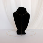 The Kara Body Chain, a delicate gold accessory perfect for summer jewelry, is shown on a black bust with a white fabric background.