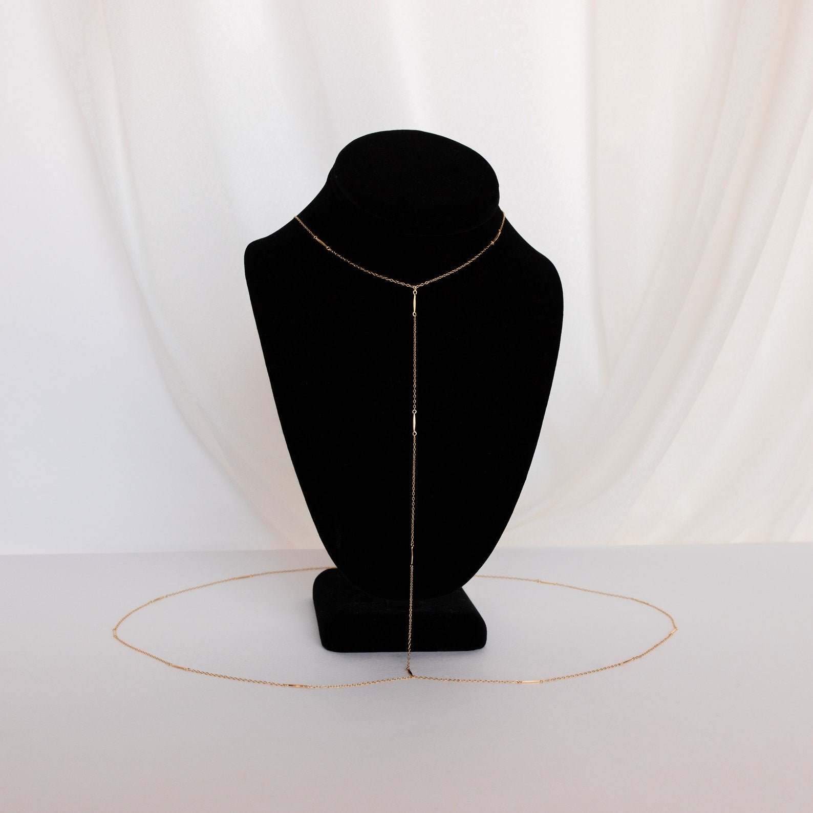 The Kara Body Chain, a delicate gold accessory perfect for summer jewelry, is shown on a black bust with a white fabric background.