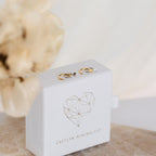 Tiny Circle & Triangle Huggies in gold and silver rest on a white jewelry box featuring a geometric heart and Caitlyn Minimalist label.
