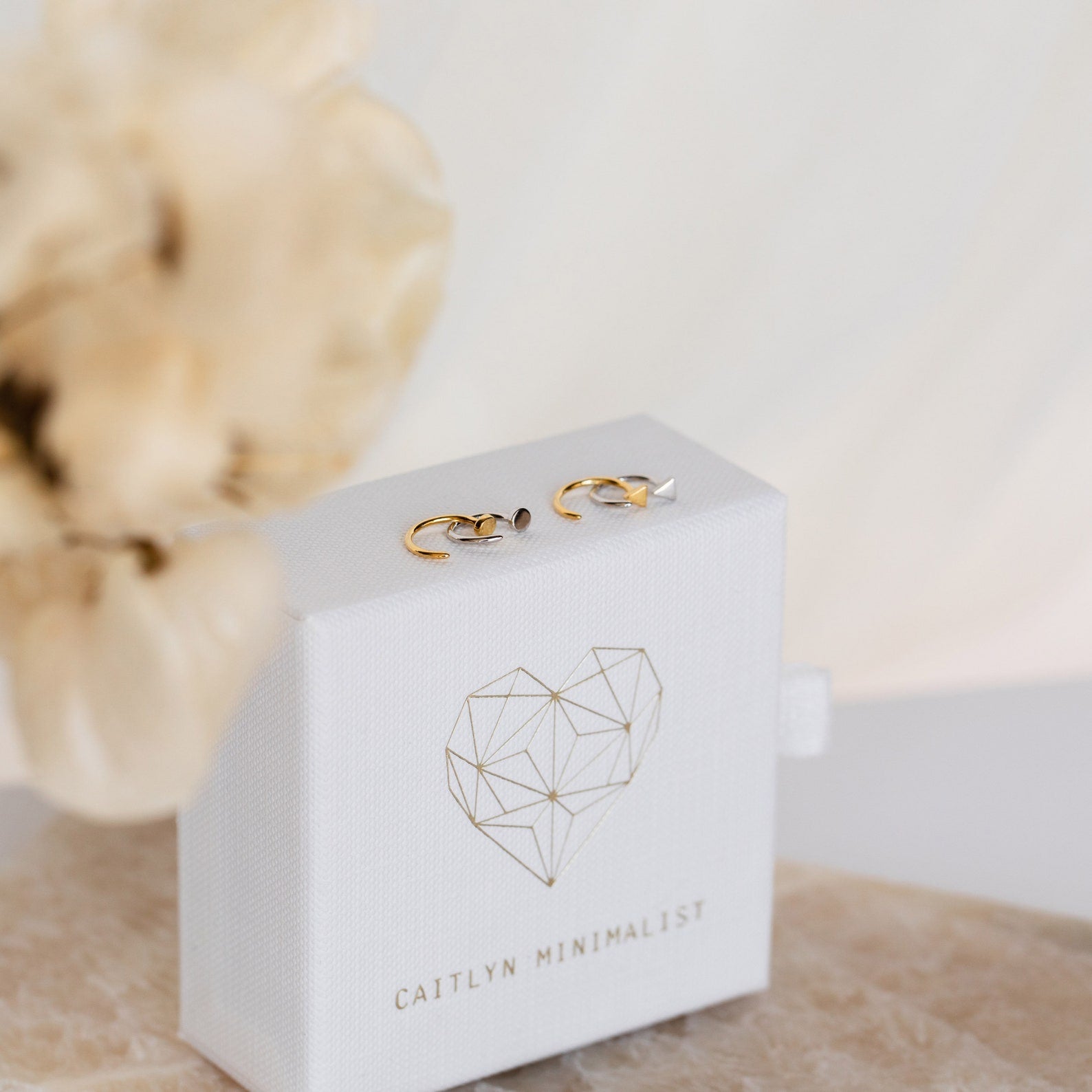 Tiny Circle & Triangle Huggies in gold and silver rest on a white jewelry box featuring a geometric heart and Caitlyn Minimalist label.
