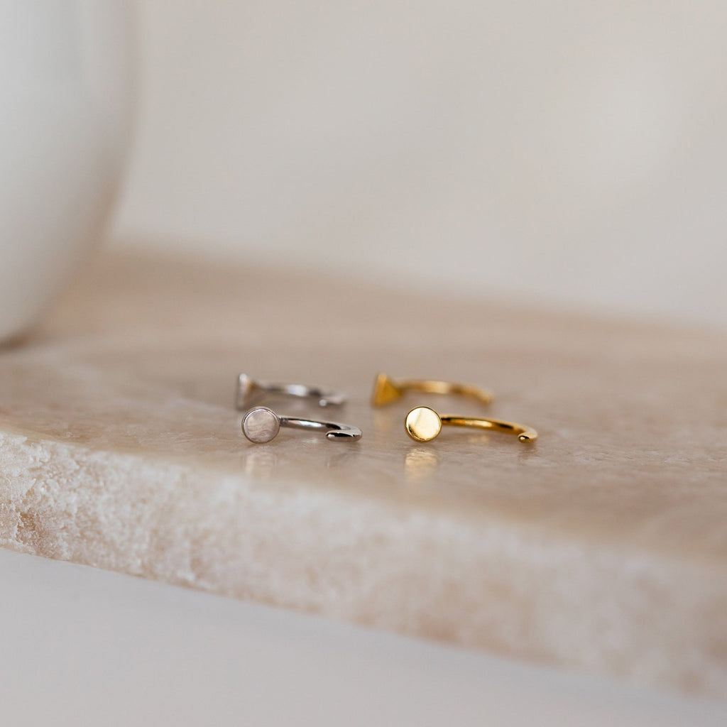 Tiny Circle & Triangle Huggies—one in silver, one in gold—displayed on beige marble. Perfect minimalist additions to your earring stack.