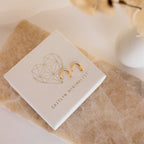The Tiny Circle & Triangle Huggies—lightweight gold minimalist earrings—are shown on a textured white jewelry box with a geometric heart and Caitlyn Minimalist text, adding chic style to any earring stack.