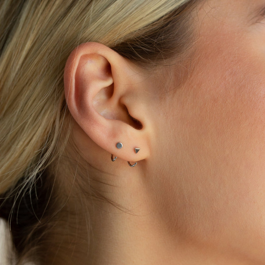 Close-up of a woman's ear adorned with the Tiny Circle & Triangle Huggies—an effortlessly chic trio featuring a silver stud, triangle stud, and small silver hoop.