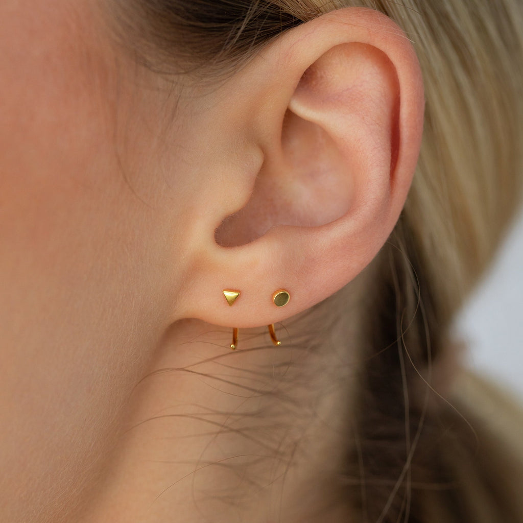 Close-up of an ear with the Tiny Circle & Triangle Huggies: three gold earrings—a triangle stud, a round stud, and a small hoop—perfect for creating a chic, lightweight layered look.