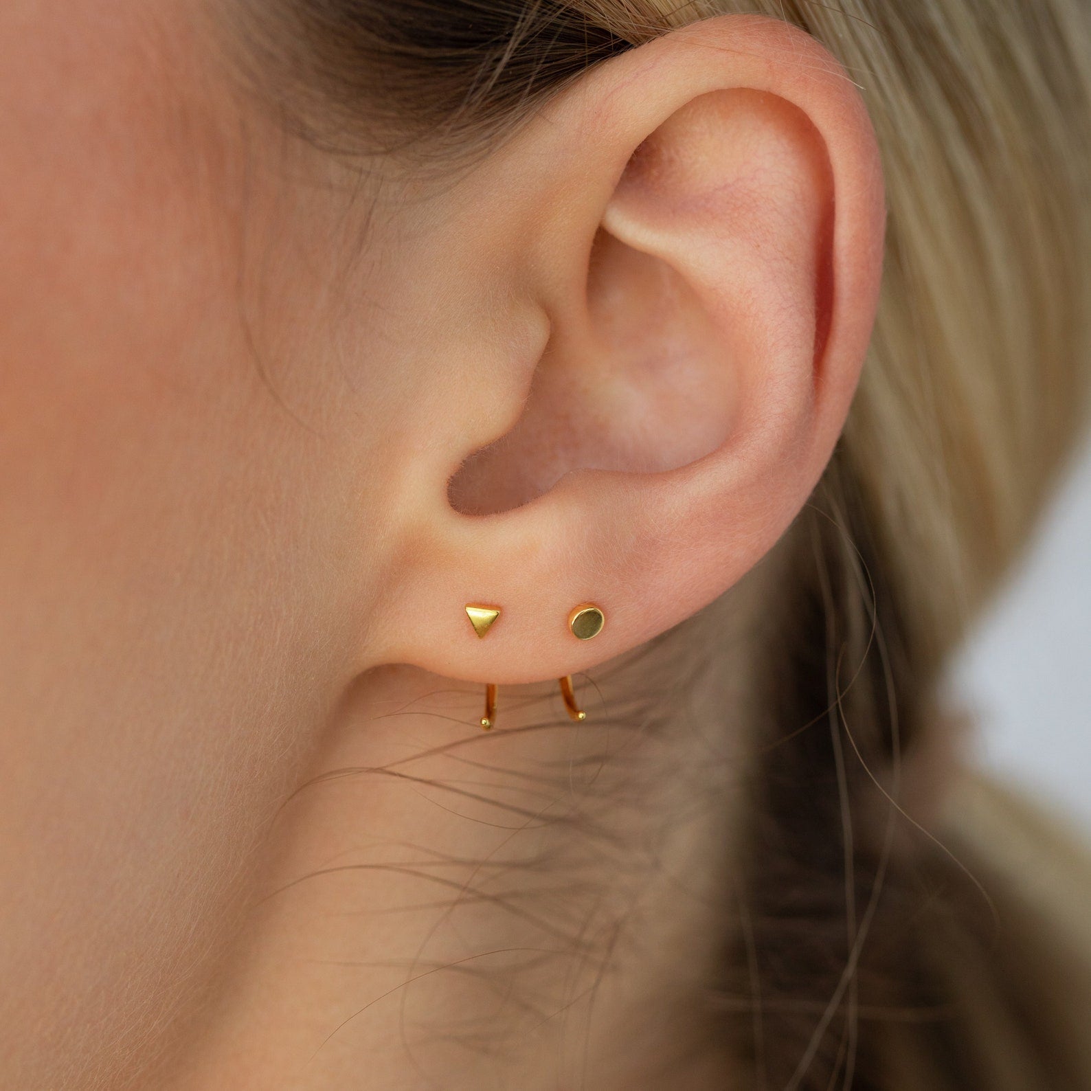 Close-up of an ear with the Tiny Circle & Triangle Huggies: three gold earrings—a triangle stud, a round stud, and a small hoop—perfect for creating a chic, lightweight layered look.