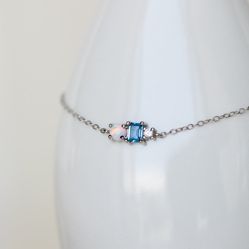 The Eloise Gemstone Bracelet showcases a blue rectangular stone and an iridescent opal on a delicate chain, ideal for dainty jewelry lovers. Elegantly displayed on a white surface.