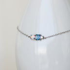 The Eloise Gemstone Bracelet showcases a blue rectangular stone and an iridescent opal on a delicate chain, ideal for dainty jewelry lovers. Elegantly displayed on a white surface.
