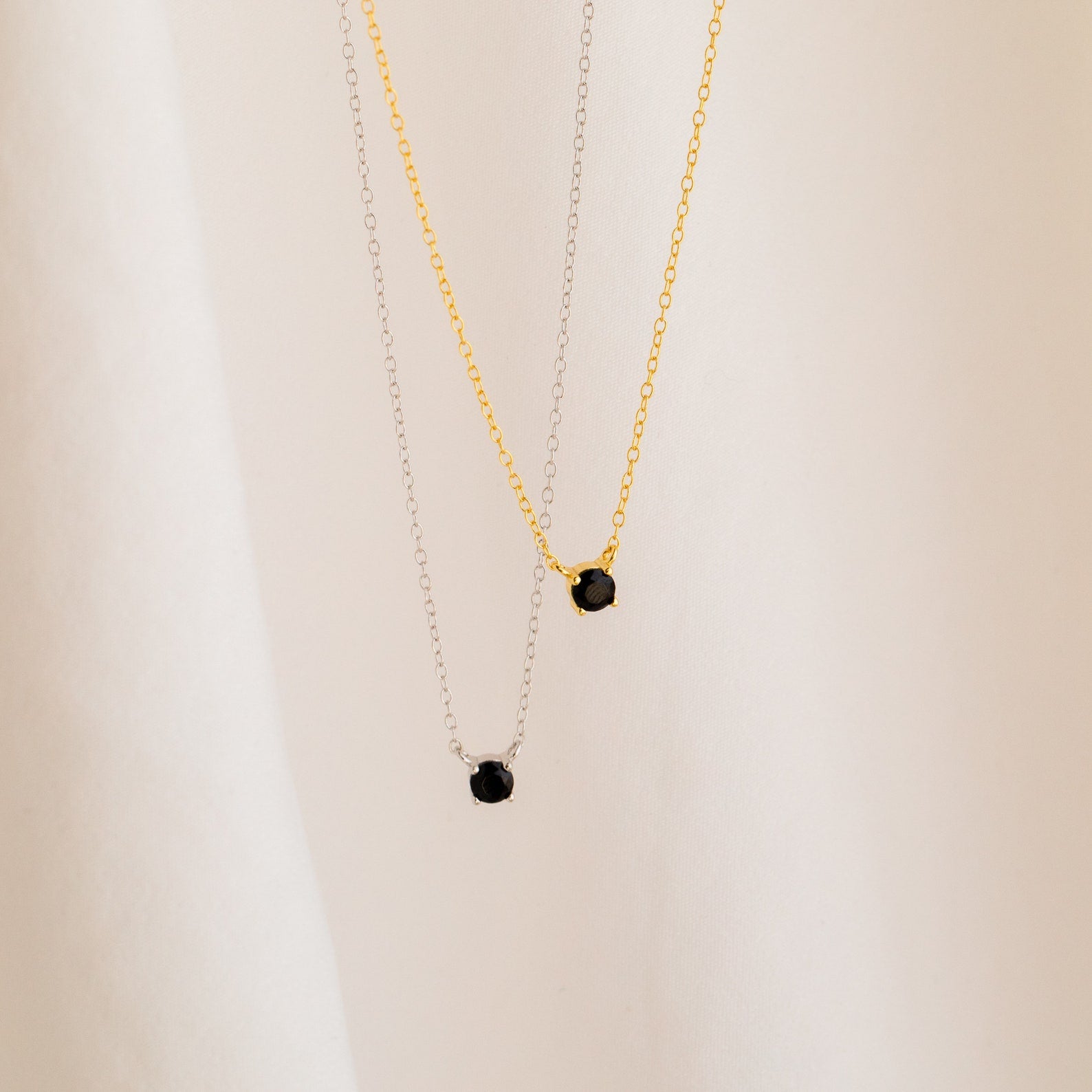 The Onyx Dainty Diamond Necklace, available in both silver and gold with a small black diamond pendant, showcases a vintage-inspired style against a soft beige background.
