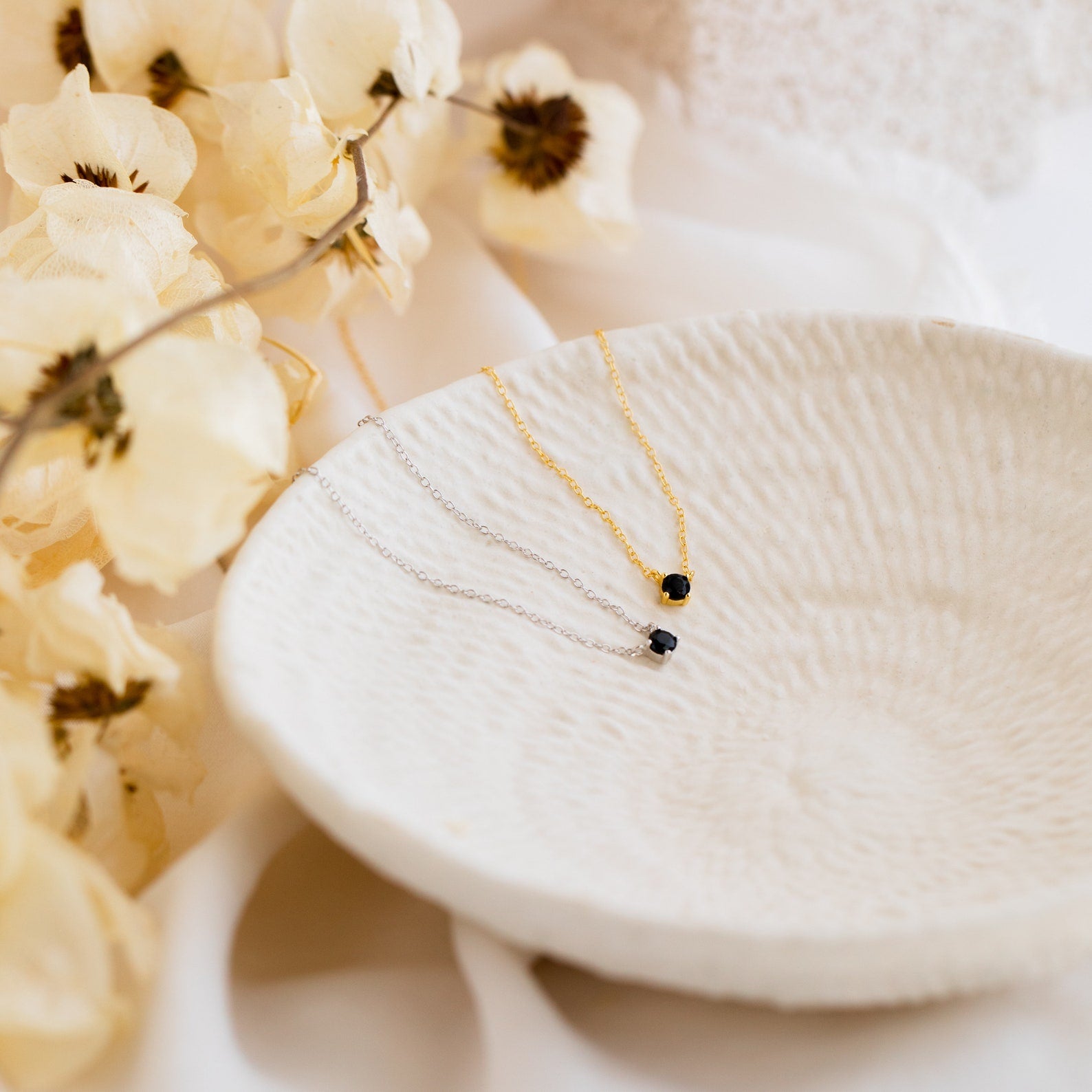 The Onyx Dainty Diamond Necklace, a delicate vintage-inspired piece with a dark pendant, rests on a textured white dish beside dried white flowers.