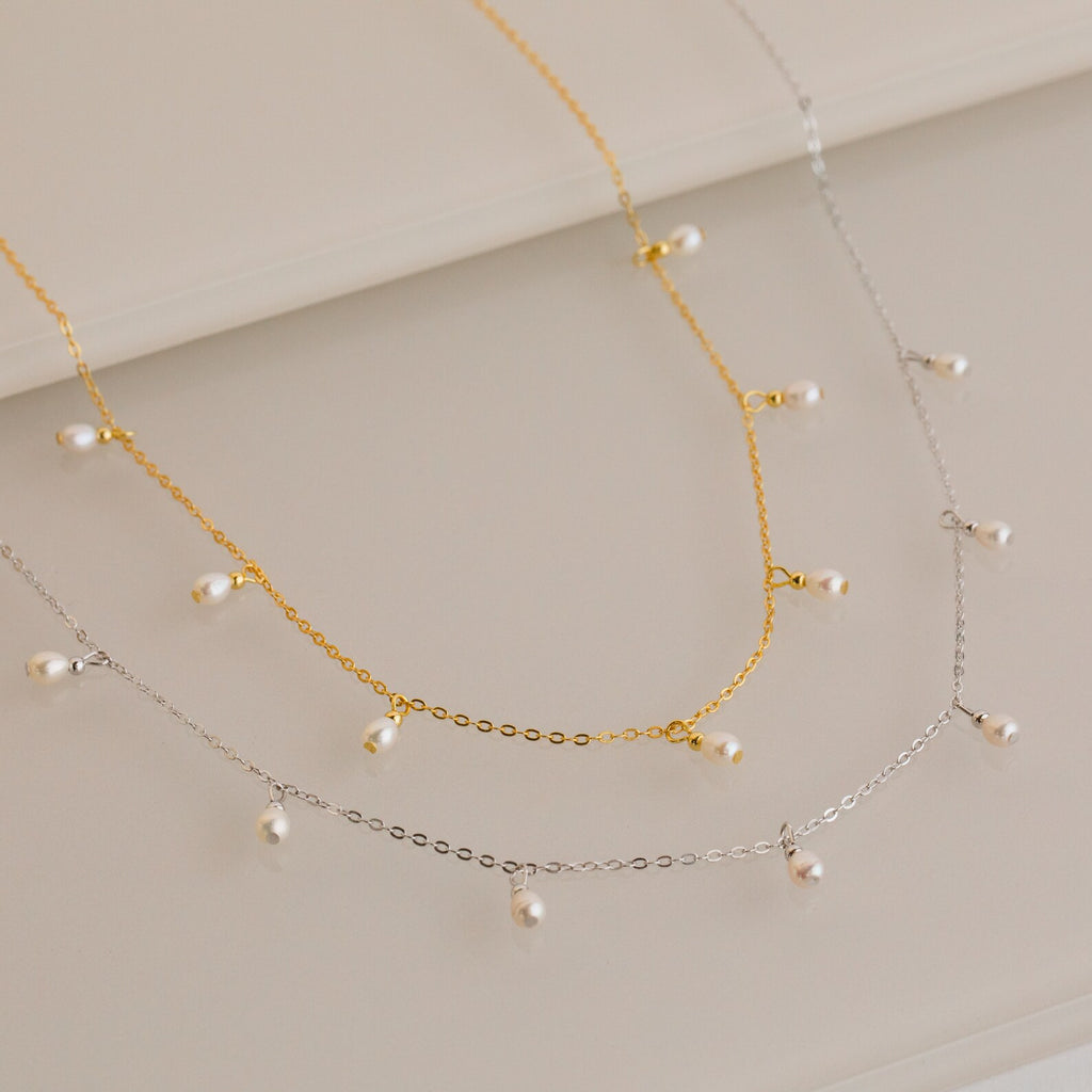 Gold and silver chain necklaces, each adorned with evenly spaced small white pearl drops, displayed on a light surface.