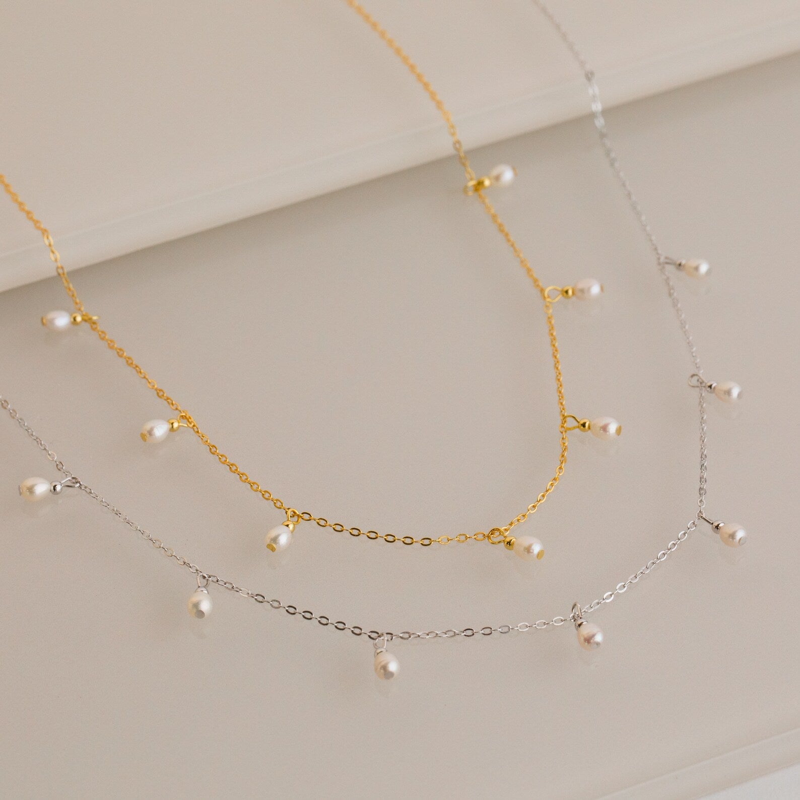 Gold and silver chain necklaces, each adorned with evenly spaced small white pearl drops, displayed on a light surface.