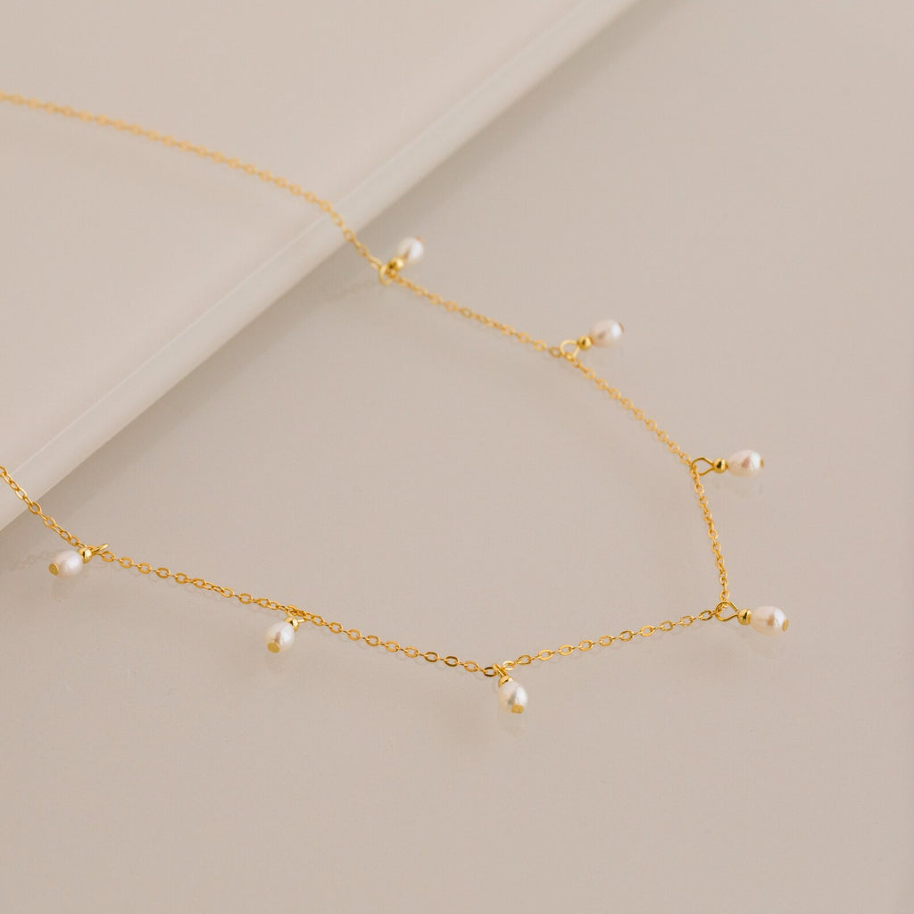 Close up of a delicate gold chain necklace with evenly spaced small white pearl drops, displayed on a smooth beige surface.