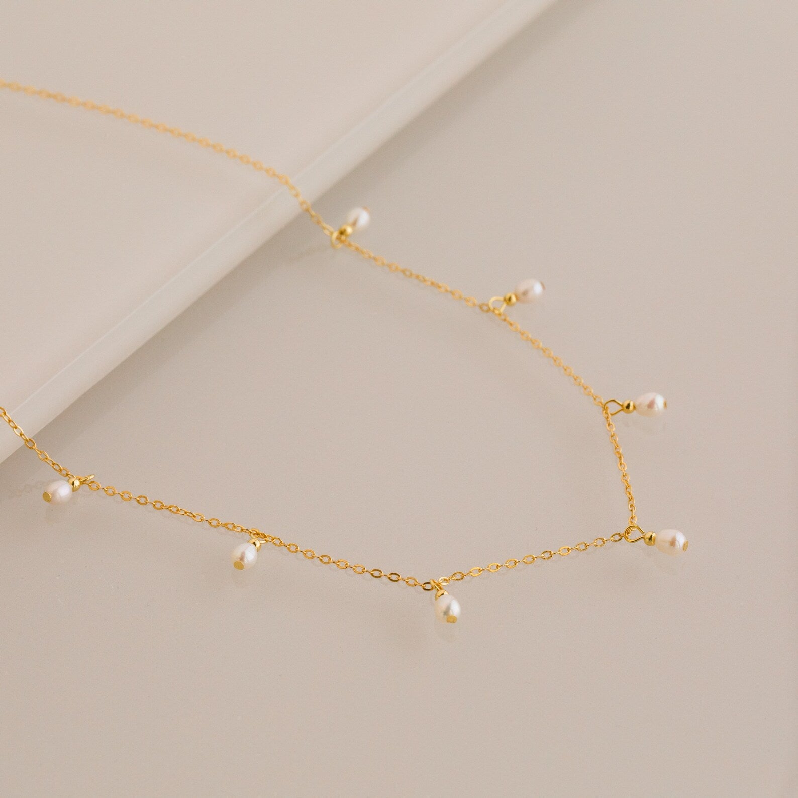 Close up of a delicate gold chain necklace with evenly spaced small white pearl drops, displayed on a smooth beige surface.