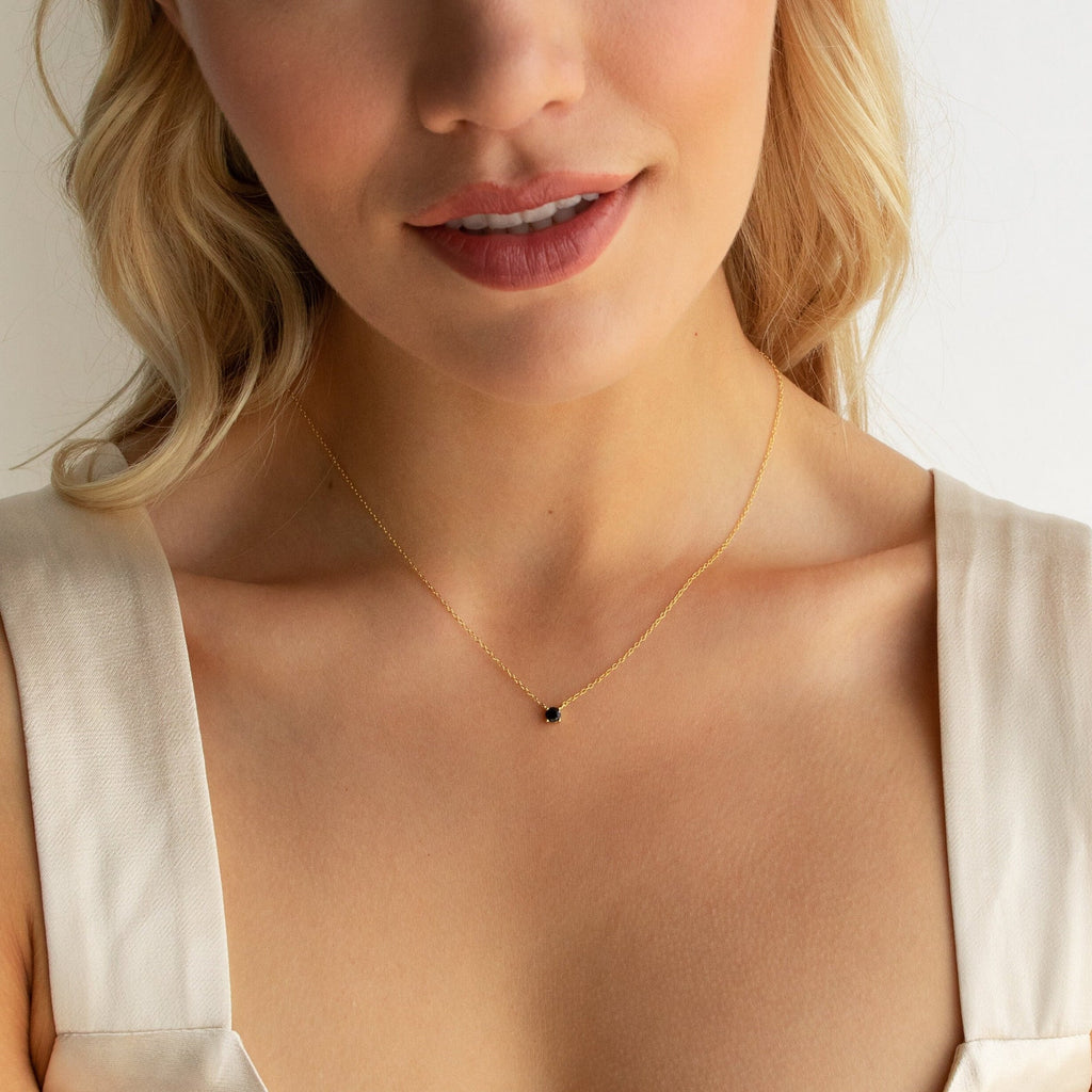 A woman elegantly pairs a cream sleeveless top with the Onyx Dainty Diamond Necklace, which features a vintage-inspired gold chain and small black pendant.