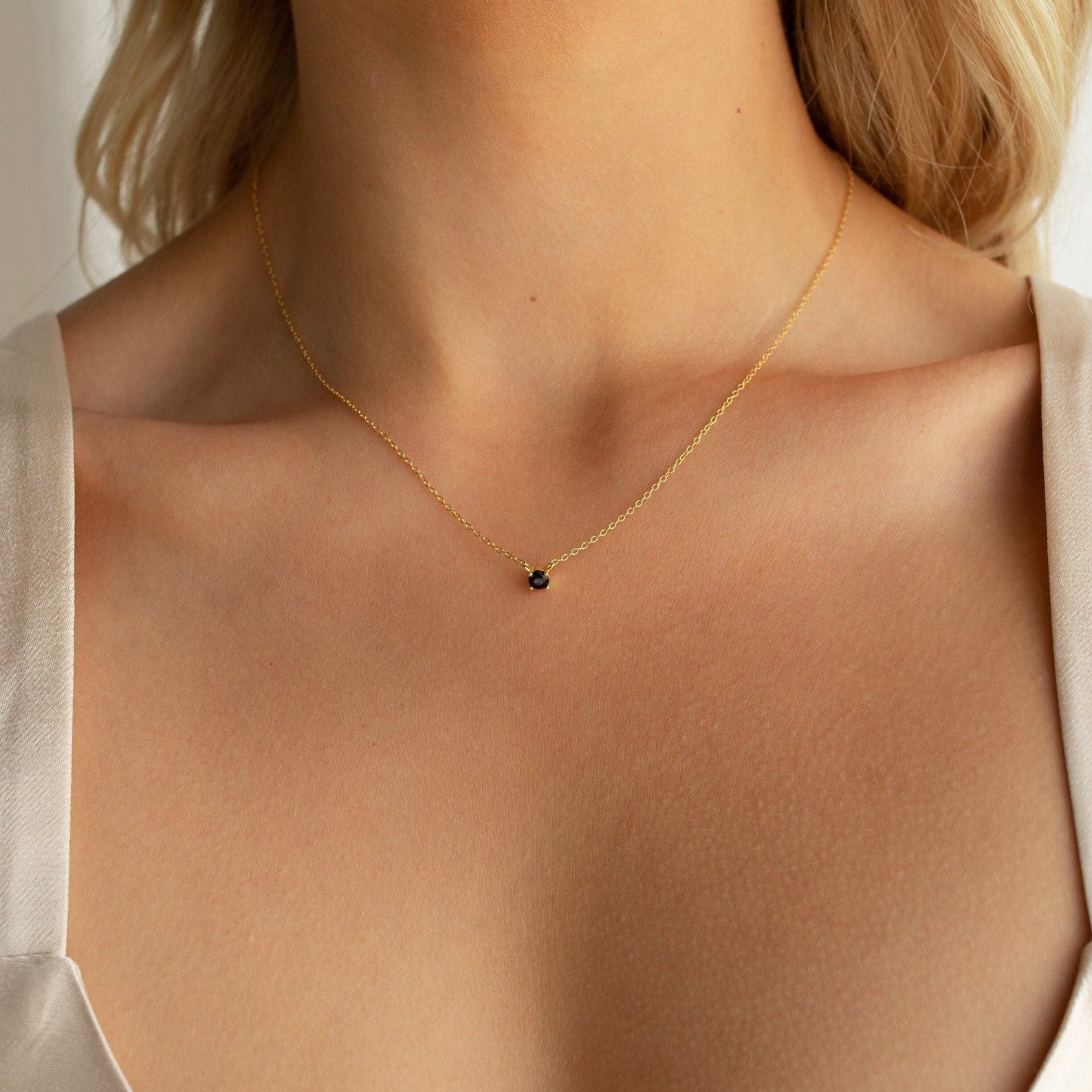 Model wears a minimalist gold chain necklace featuring a small round black onyx stone set in a dainty prong setting at the center of the collarbone.