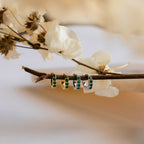 Four Elise Emerald Huggies in gold and silver, some accented with emerald stones, are displayed on a branch with dried white flowers in the background for an elegant green jewelry showcase.