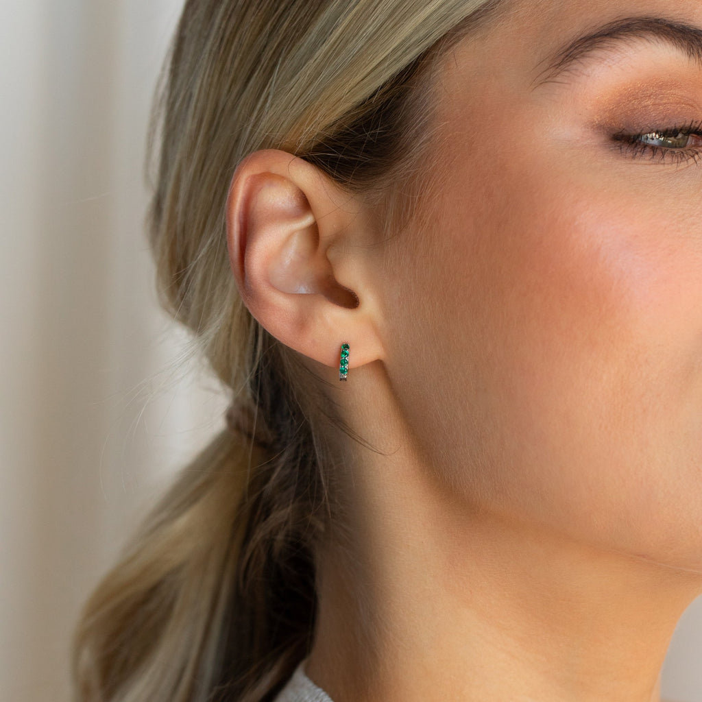 Close-up of a woman's ear wearing the Elise Emerald Huggies, small gold earrings adorned with emerald stones.