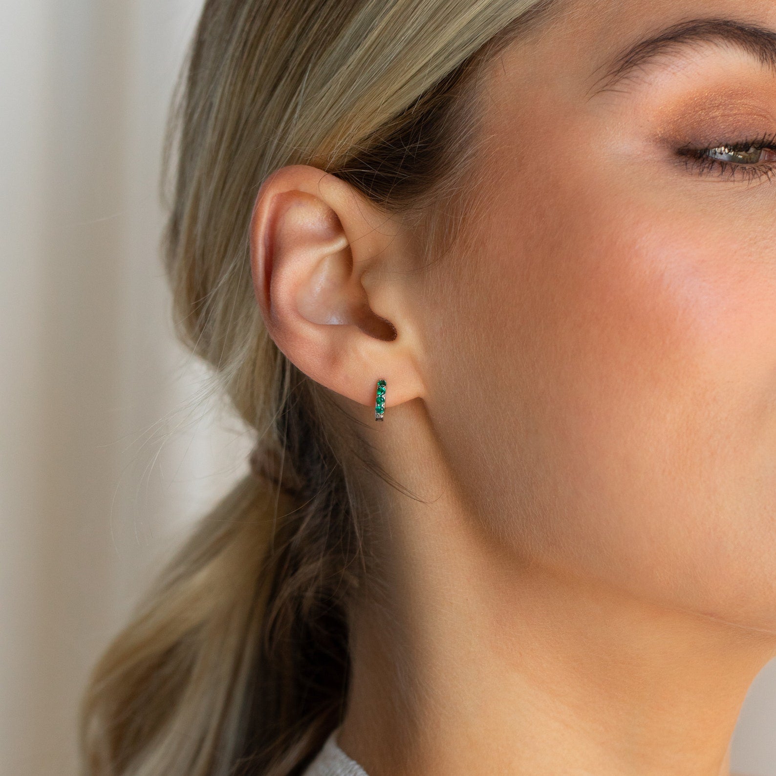 Close-up of a woman's ear wearing the Elise Emerald Huggies, small gold earrings adorned with emerald stones.