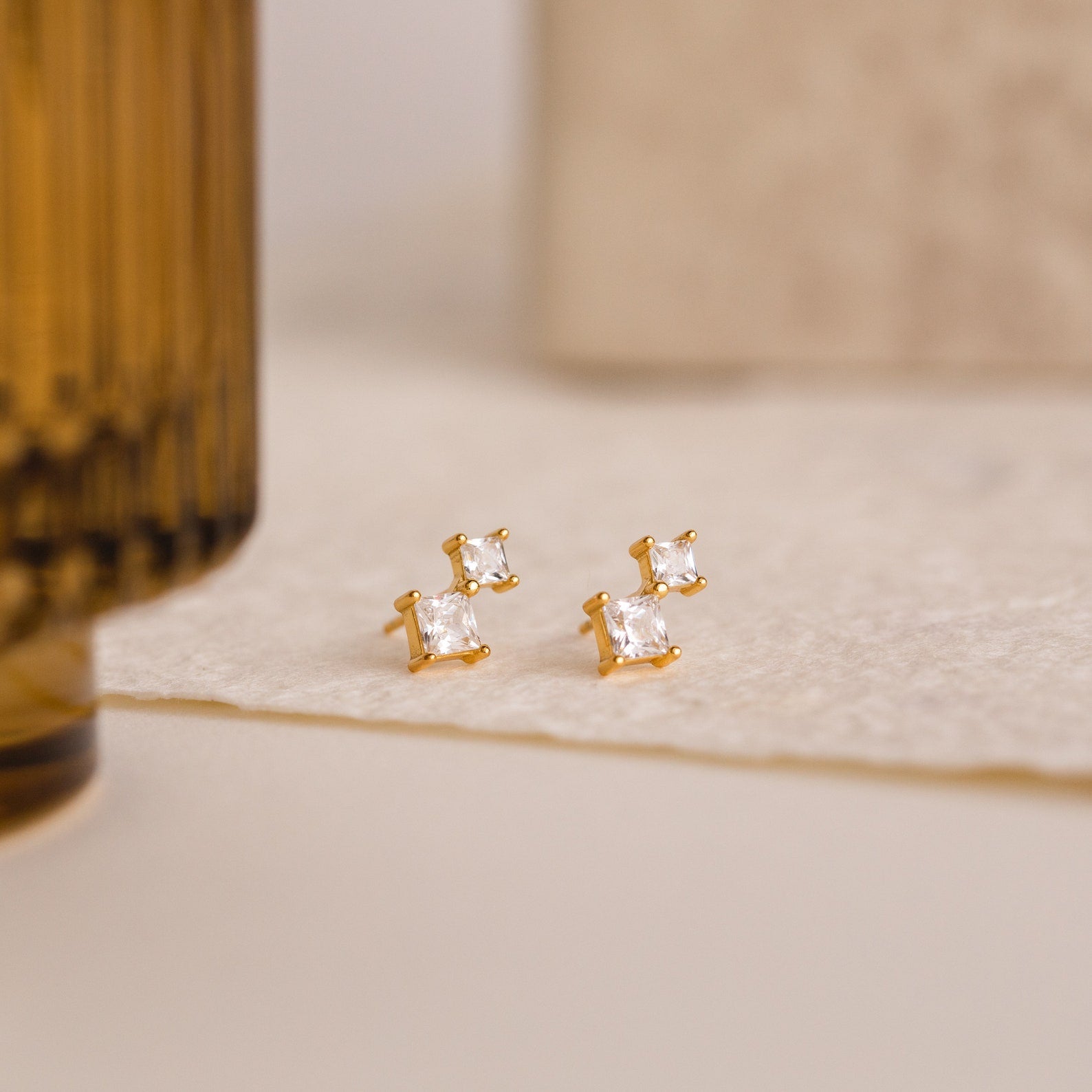 Aria Diamond Drop Studs: gold crystal earrings featuring two square, clear gemstones on a textured cream surface.