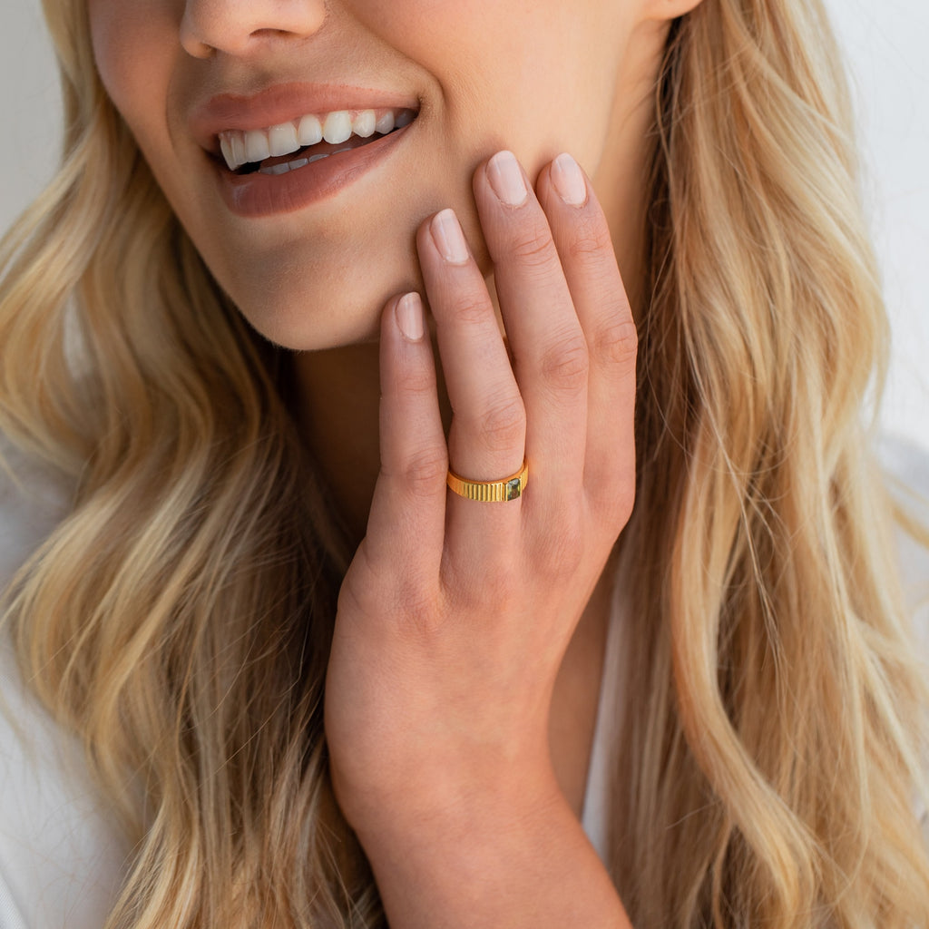 Woman smiling with long blonde hair, wearing the Olive Peridot Signet Ring and touching her face with manicured nails.