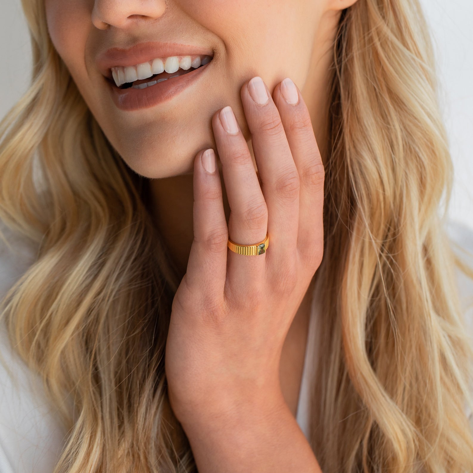 Woman smiling with long blonde hair, wearing the Olive Peridot Signet Ring and touching her face with manicured nails.