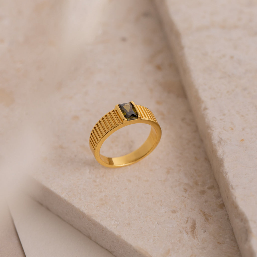The Olive Peridot Signet Ring features a gold chunky band with a rectangular dark green gemstone, elegantly displayed on a textured, light-colored stone surface.